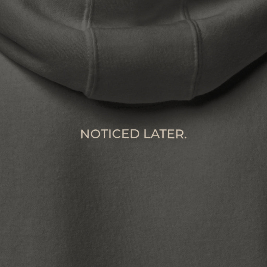 Close-up of the back of the Noticed Later Hoodie, NOTICED LATER. printed in sand under the hood.