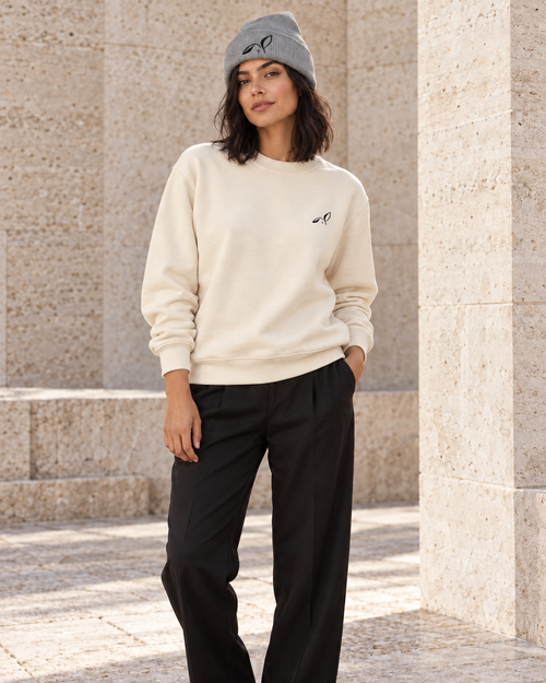 Woman in a bone crewneck and heather grey beanie, both with the black LOUPI GANG rabbit icon, standing among pale stone columns.