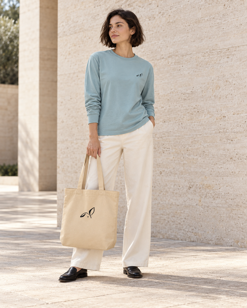 Woman in an agave long sleeve tee carrying a natural organic tote, both with the black LOUPI GANG rabbit icon, in a sunlit stone courtyard.